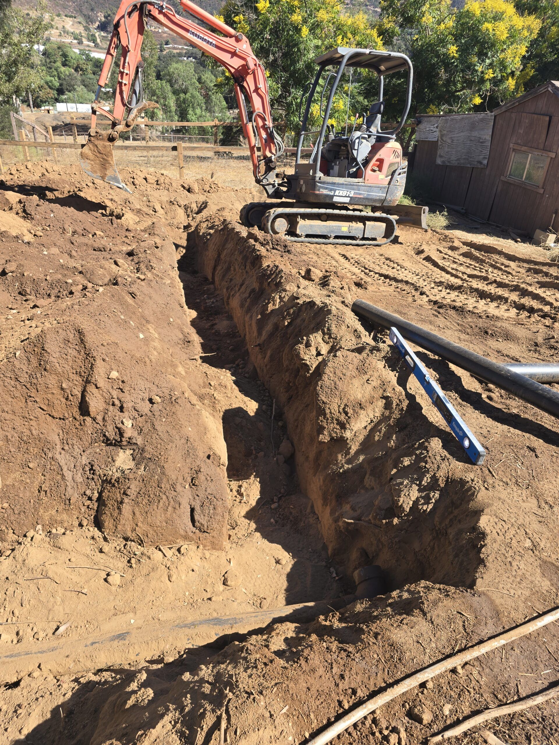An orange mini-excavator digs a deep trench in a dirt field next to a pipe and a small brick structure.