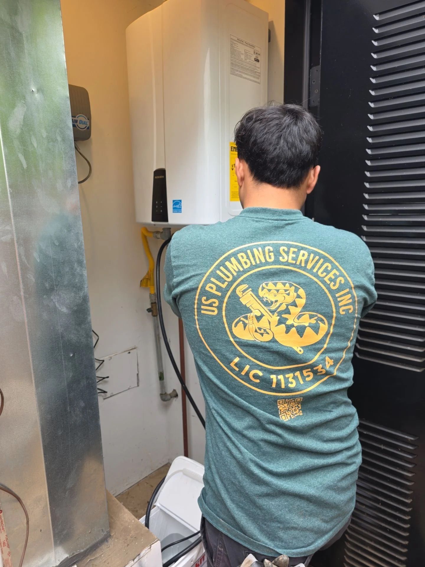 A plumber in a green work shirt stands in a mechanical closet working on a wall-mounted tankless water heater.
