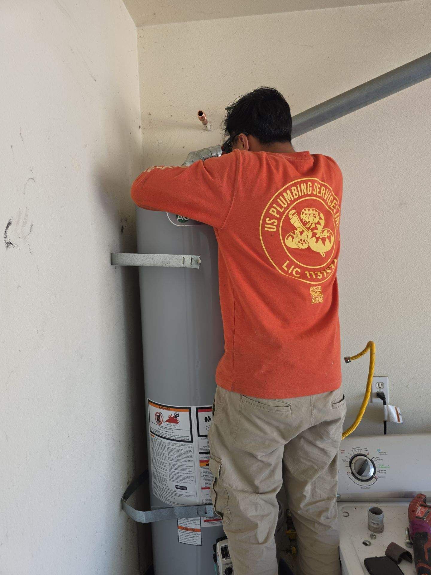 A person in an orange long-sleeved shirt works on installing or repairing a water heater in a residential laundry area.