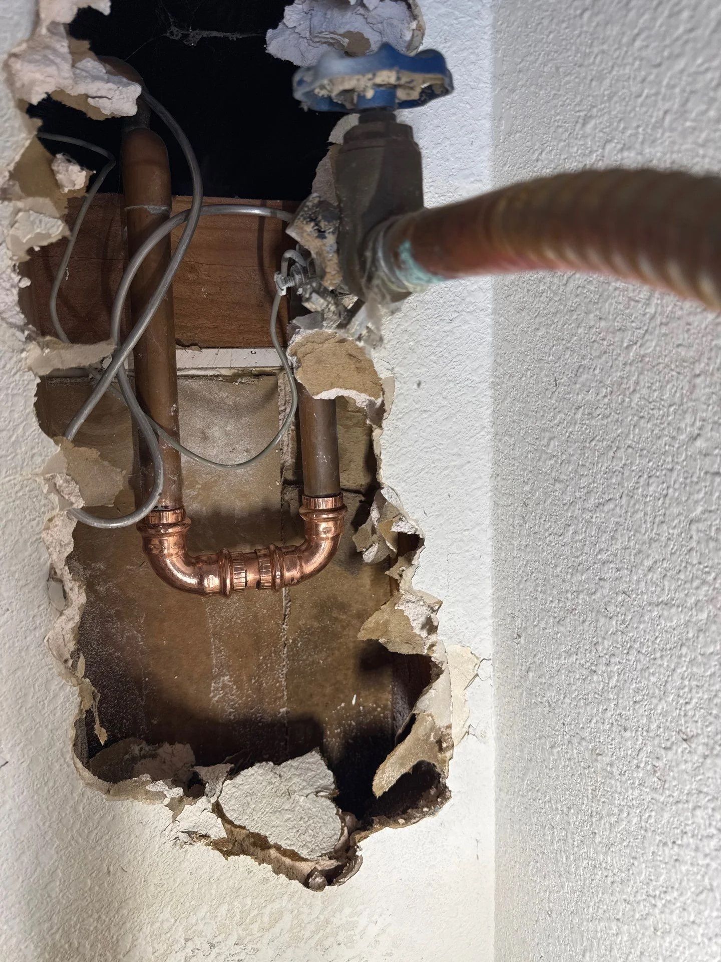 A hole in a wall revealing copper plumbing pipes connected by a U-shaped pipe fitting near a shut-off valve.