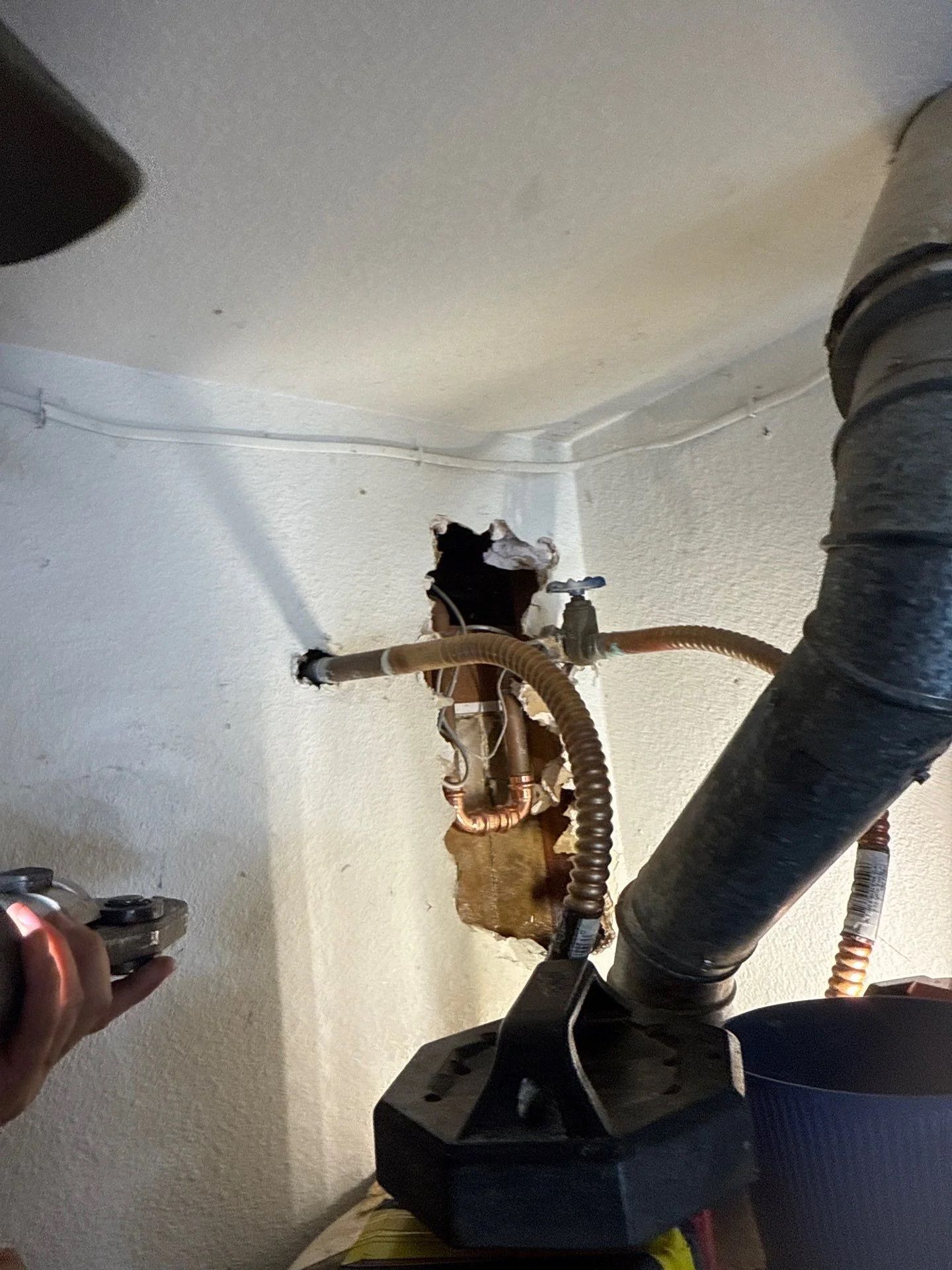 A close-up of a damaged plaster wall with exposed water pipes and a valve, beside a dark metal flue pipe.