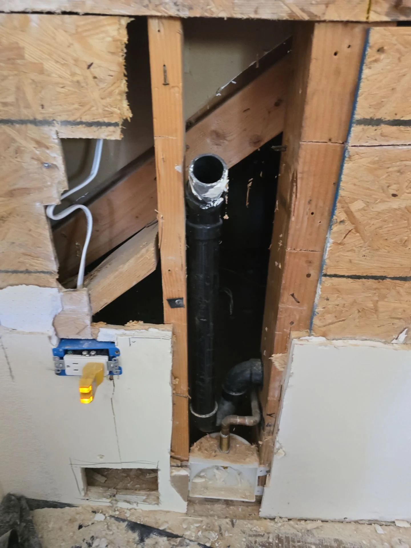 A wall section opened to reveal black ABS plumbing pipes and wooden framing with a yellow electrical outlet nearby.