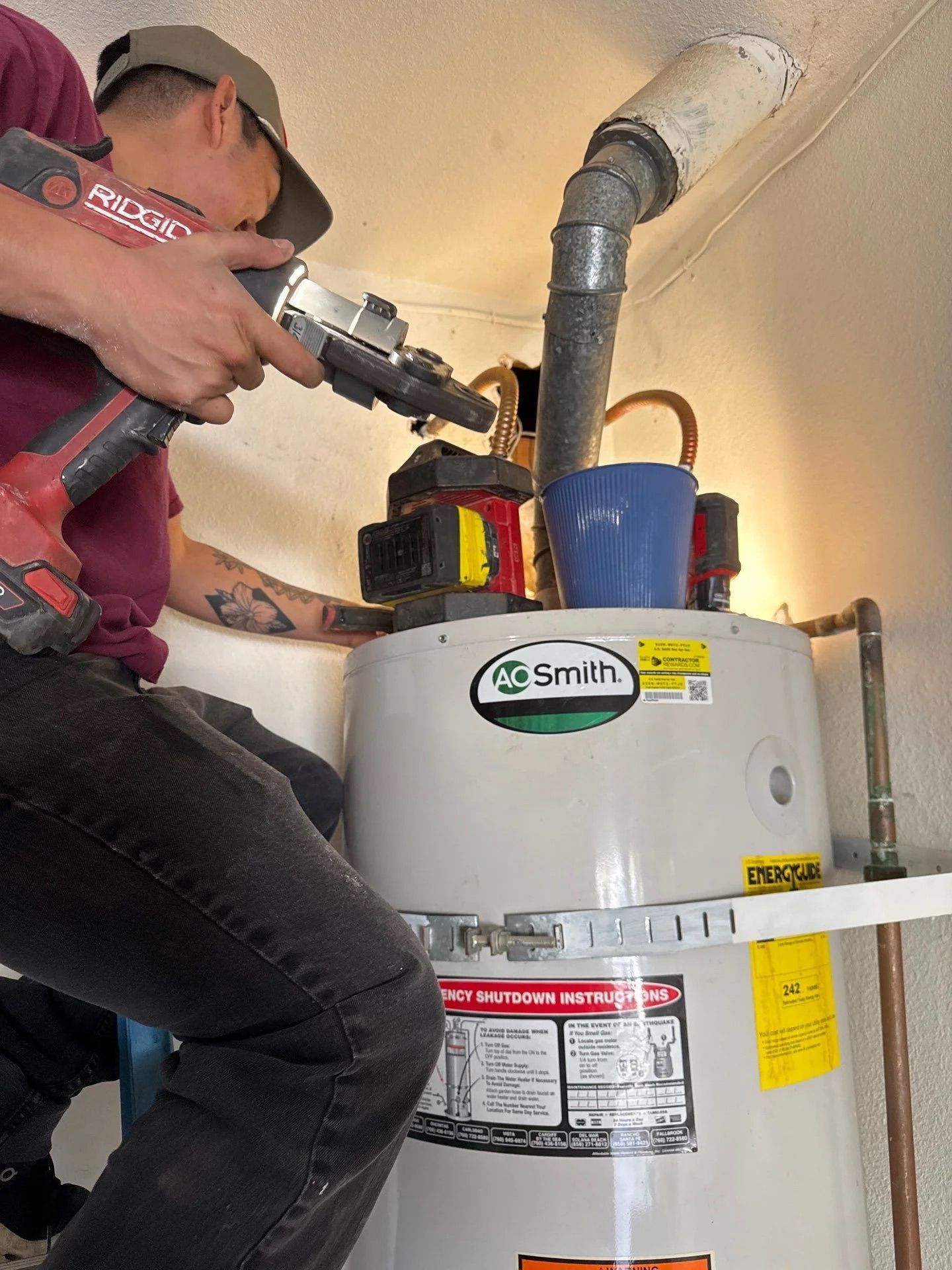 A technician in a maroon shirt uses a cordless reciprocating saw to cut a metal vent pipe above an A. O. Smith water heater.