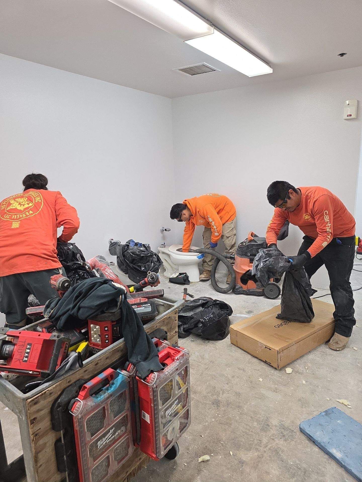 Three workers in orange uniforms clean a room, using equipment and black trash bags.