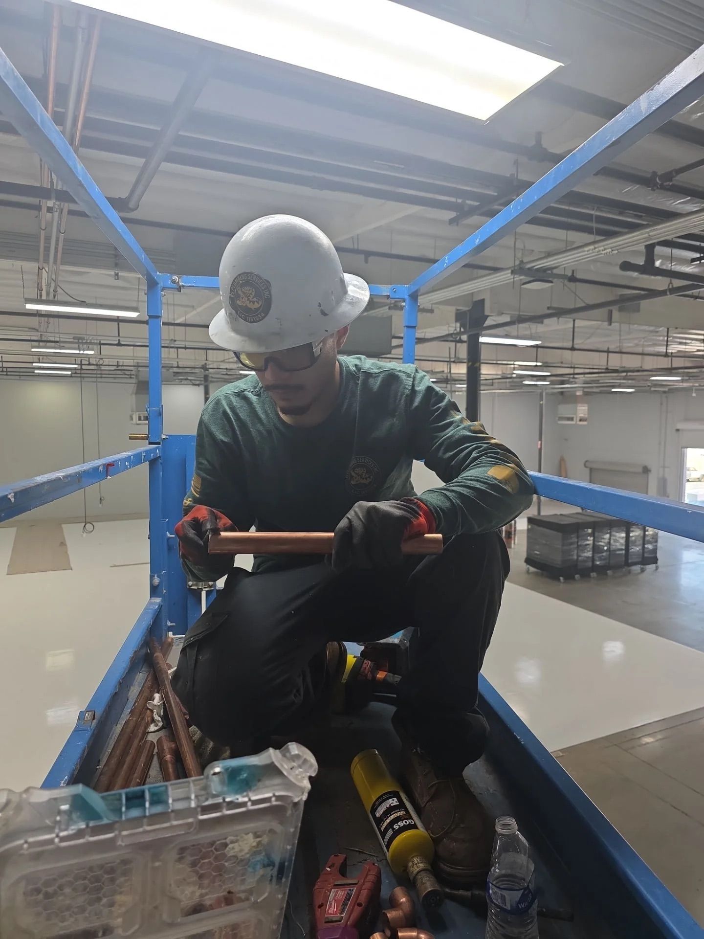 A worker wearing a hard hat and safety glasses inspects a copper pipe while kneeling on a construction lift platform.