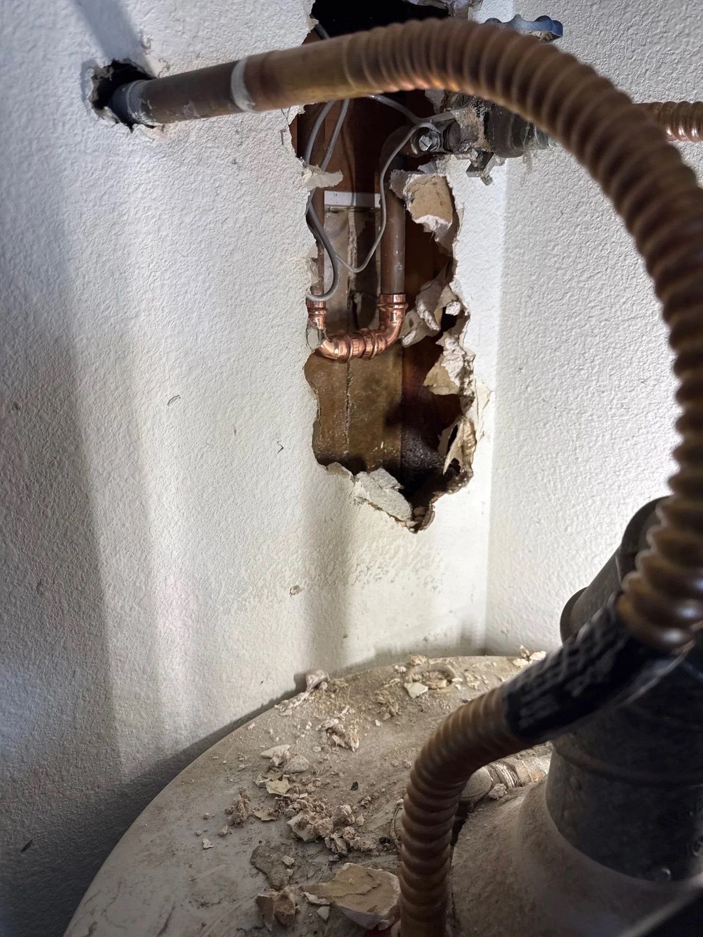 A hole in a wall revealing copper plumbing pipes above a water heater with flexible, corrugated supply lines.