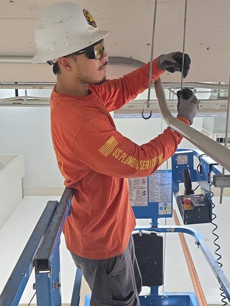 A worker in an orange long-sleeved shirt and hard hat installs ceiling hardware from an elevated platform.