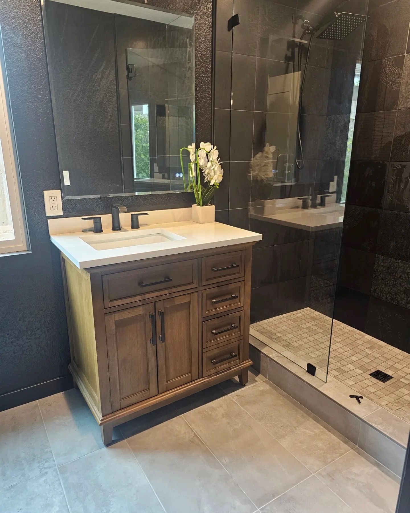 A modern bathroom vanity with a wood finish, white countertop, and mirror next to a glass-enclosed shower with dark tiles.