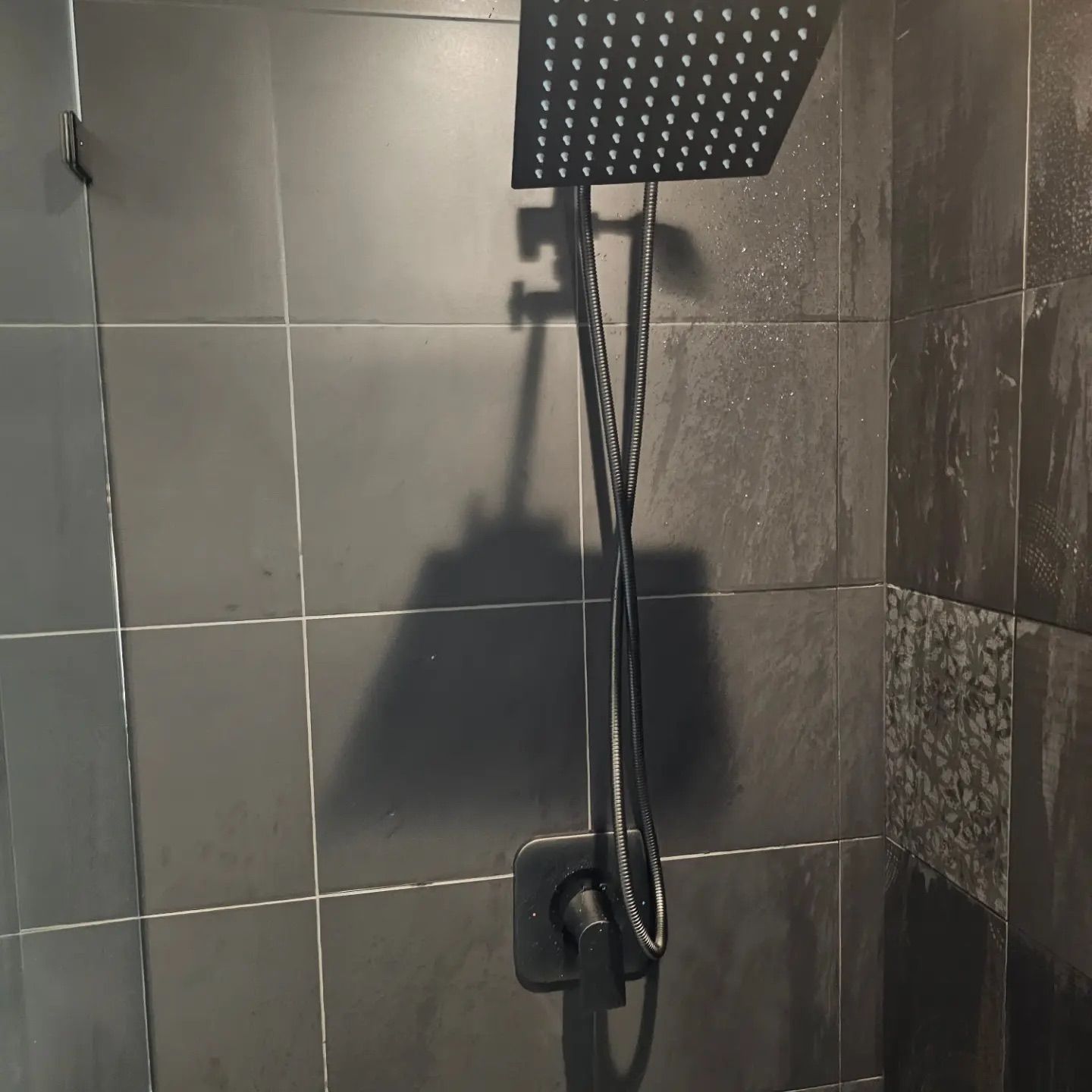 A matte black square rain shower head with a matching handheld wand and wall-mounted control valve against dark tiles.