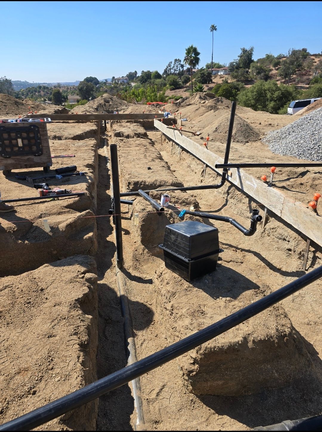 Construction site showing an open dirt trench with exposed black plumbing pipes and a small utility box in the ground.
