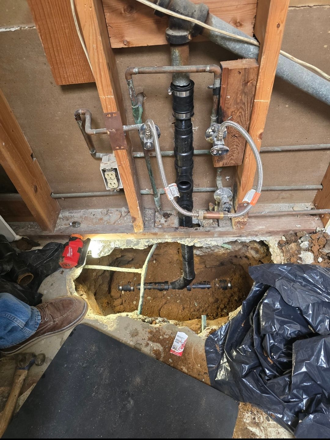 An open wall cavity showing plumbing pipes connecting to a black sewer line leading into a hole broken into the floor.