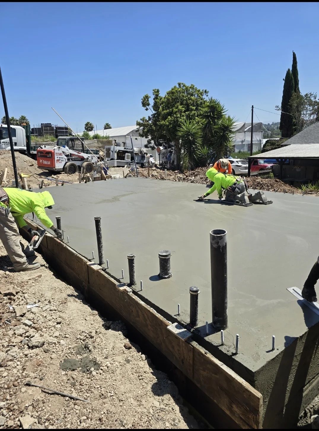 Construction workers in high-visibility vests smooth wet concrete for a new building foundation under a clear blue sky.