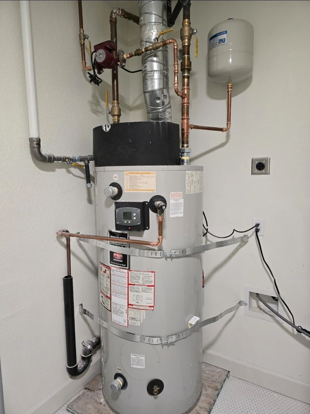 A gray water heater with attached copper pipes, a pump, and a small expansion tank, set against a plain white wall.