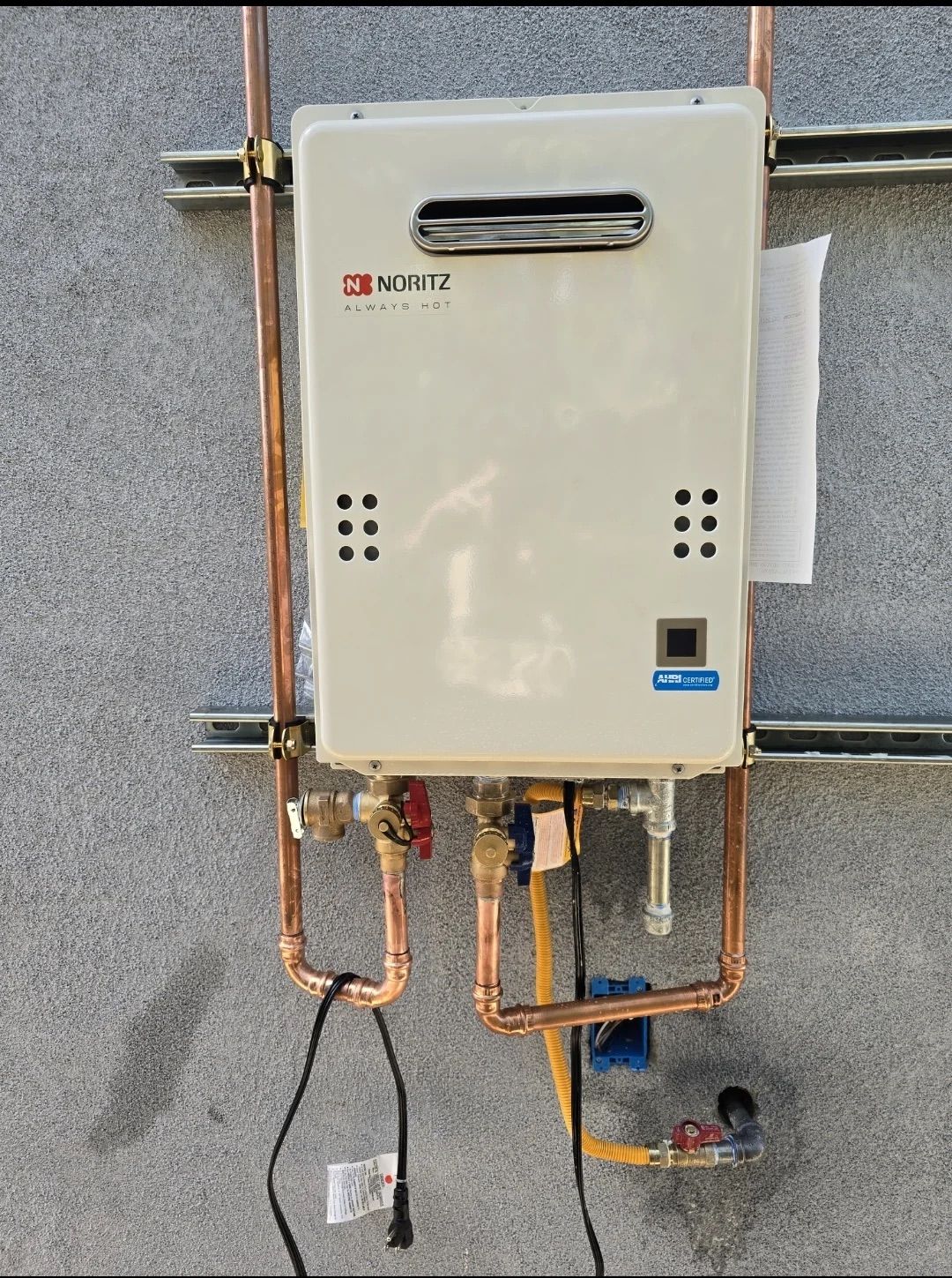 A wall-mounted Noritz tankless water heater installed outdoors on a gray stucco wall with copper plumbing connections.