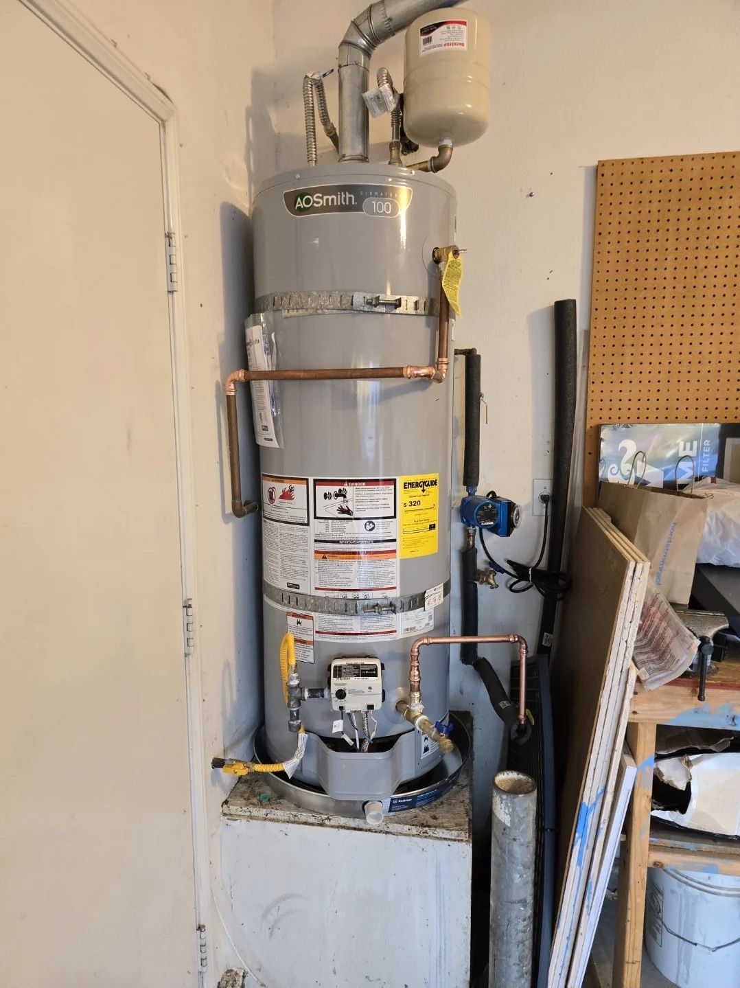 A grey gas water heater with an attached expansion tank, standing on a raised platform in a garage setting.
