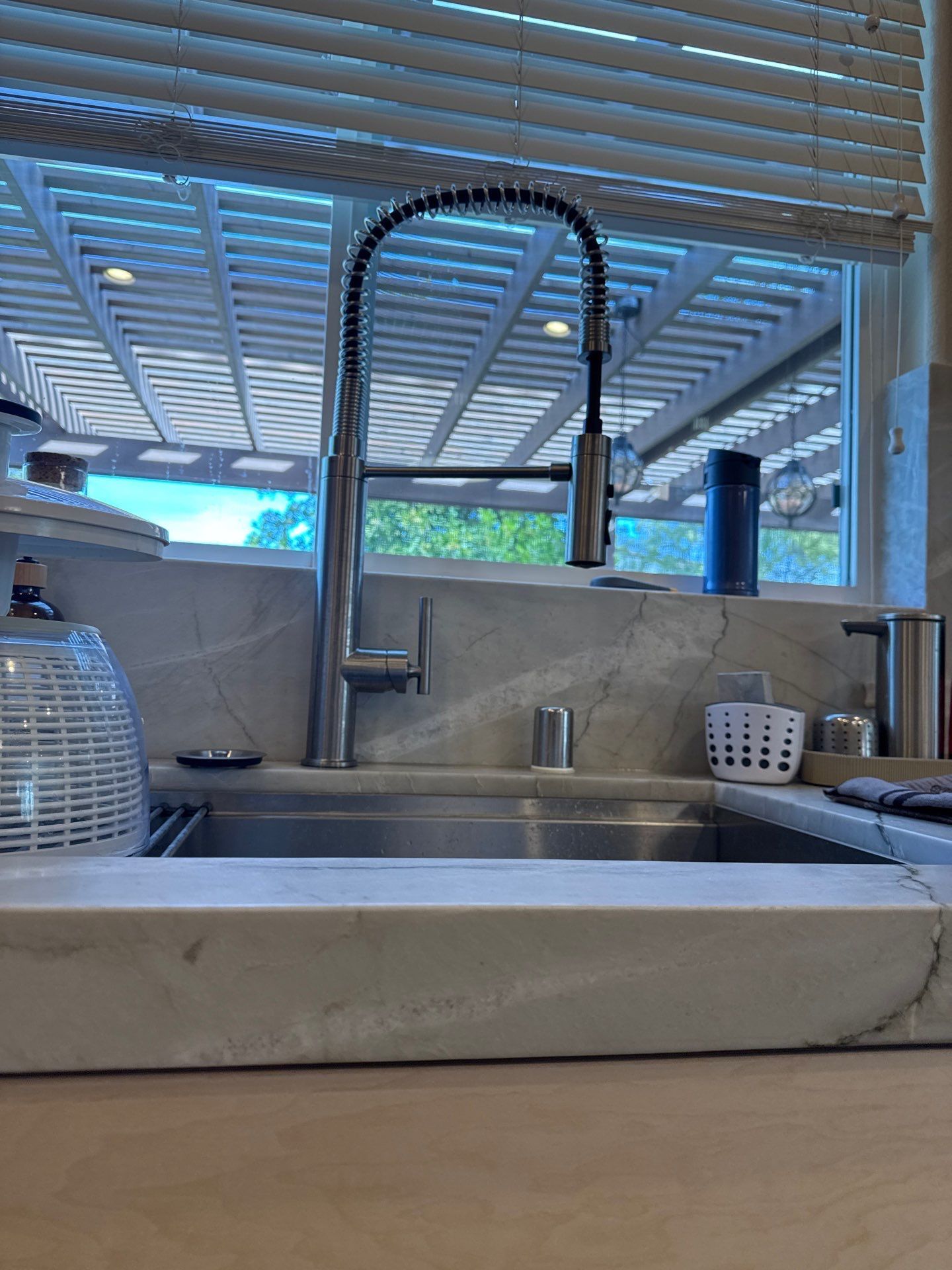A kitchen sink area featuring a stainless steel pull-down faucet set against a marble backsplash and window with blinds.