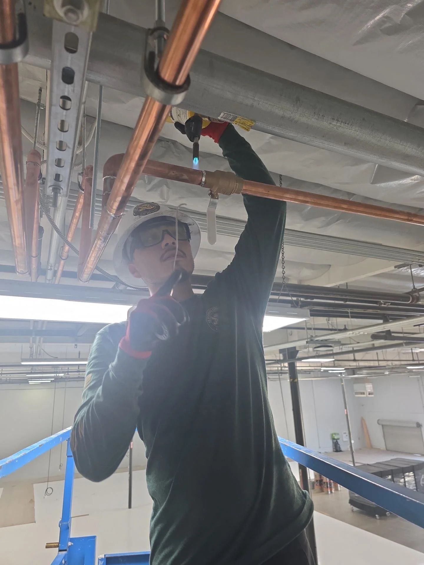 A worker in a hard hat and safety glasses uses a blowtorch to solder copper piping on a construction site.