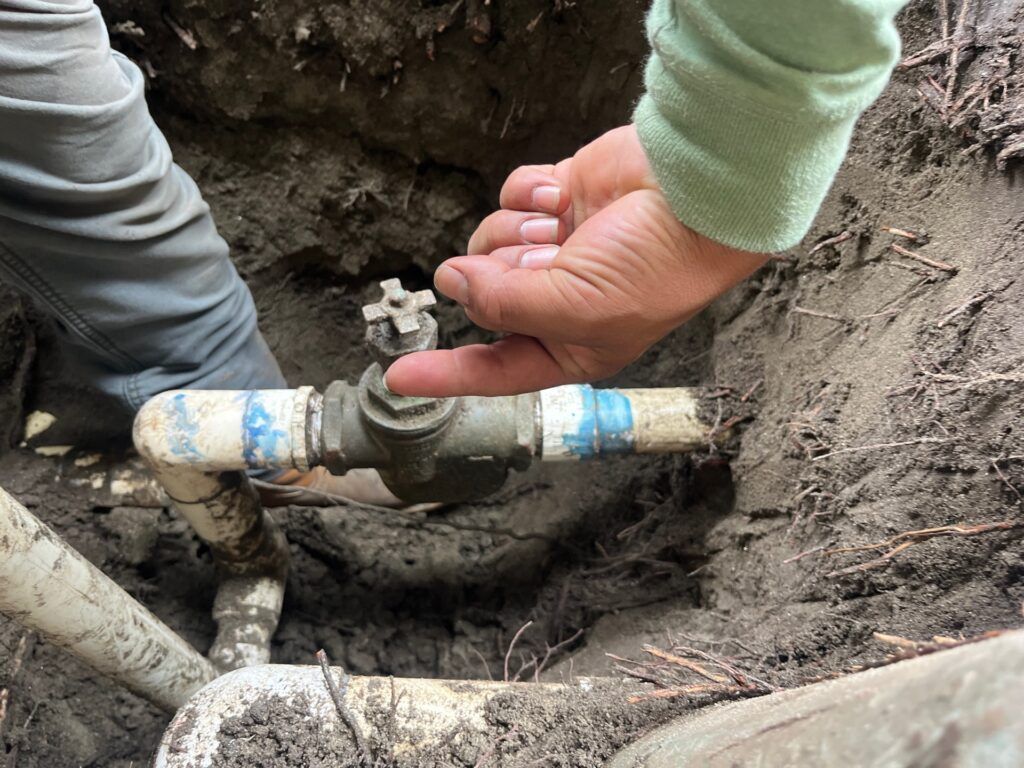 A person’s finger points to a brass shut-off valve on a section of white PVC pipe buried in a dirt trench.