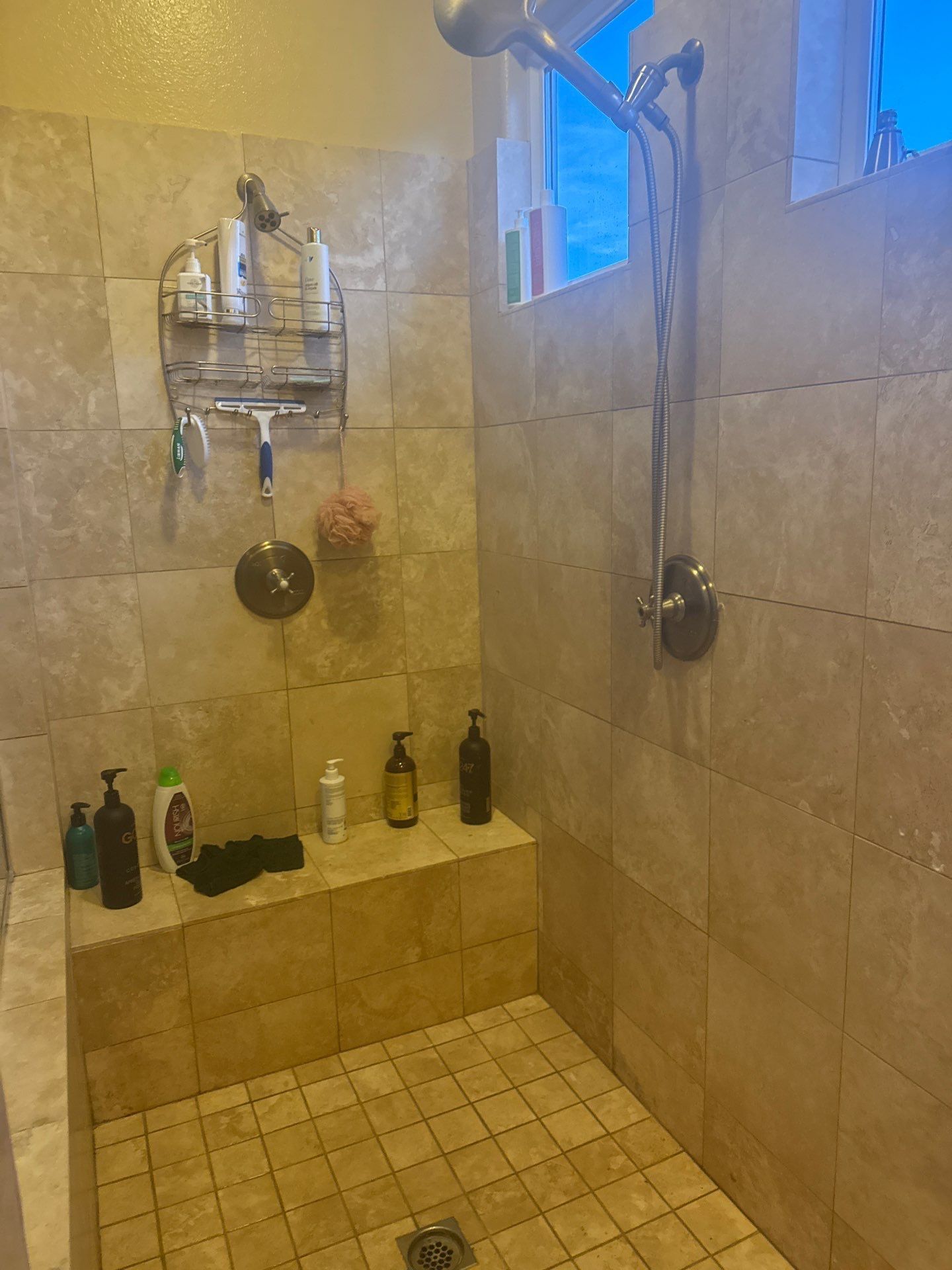 A walk-in shower with tan tiled walls and flooring, a built-in stone bench, a metal shower caddy, and a dual-head shower.