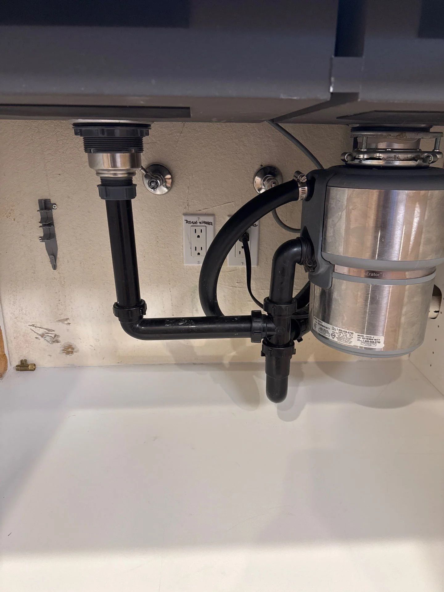 Under-sink view of a kitchen drain setup with a disposal unit and black PVC piping against a beige cabinet wall.