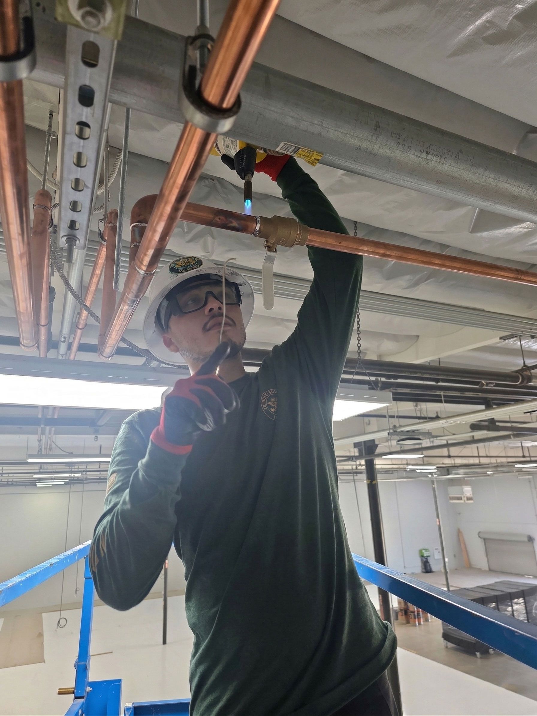 A worker in a hard hat and safety gear uses a torch to solder copper pipes near a ceiling in an unfinished building.