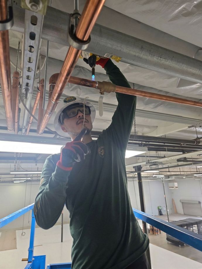A worker wearing a hard hat and protective glasses uses a torch to solder copper piping overhead from a scissor lift.