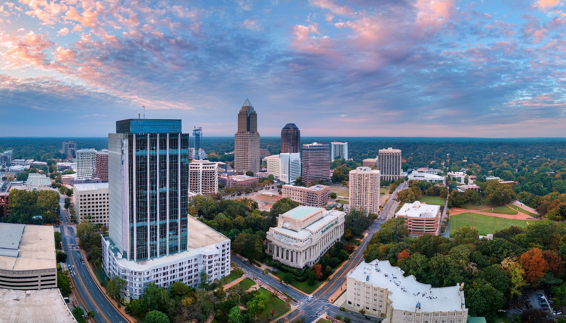 Downtown Winston-Salem, NC, skyline at sunset; high-rise buildings, colorful sky, lush green trees.