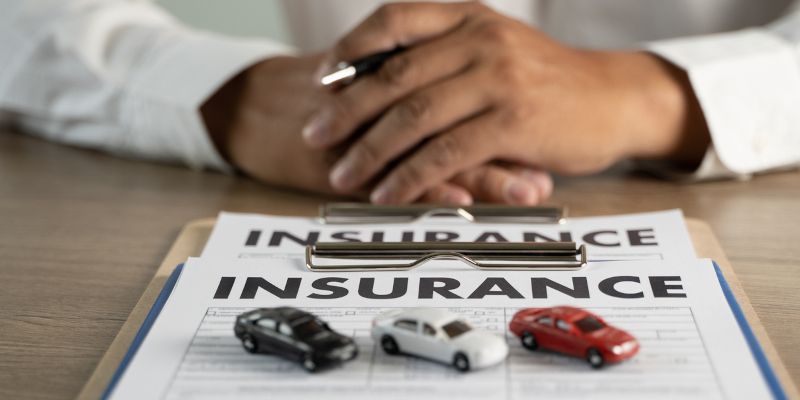 Close-up of insurance document with toy cars on a wooden table. A person's hands are visible holding a pen.