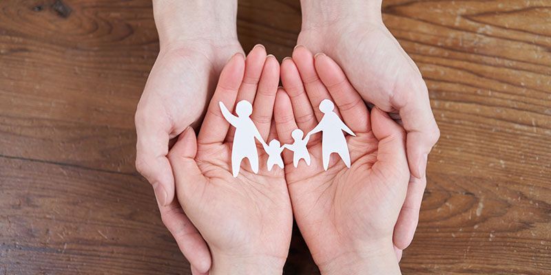 Two hands cupping a paper cutout family on a wooden surface.