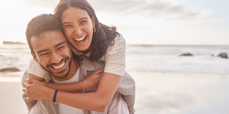 Man gives woman a piggyback ride on the beach. Both smile broadly, with ocean in background.