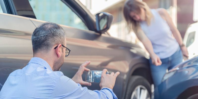 Man photographing a car with a smartphone, next to a woman examining damage from an accident.