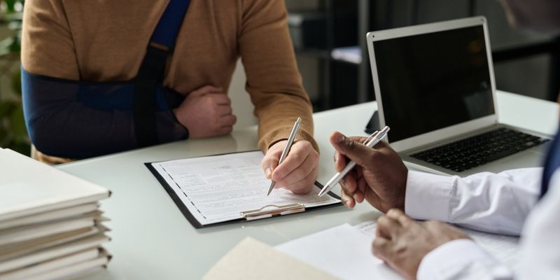 Person with arm in sling signing paperwork with another person at a desk.