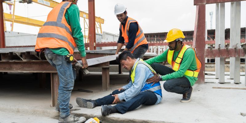 Construction workers helping an injured colleague. One man sits on ground holding his knee, other workers provide support.