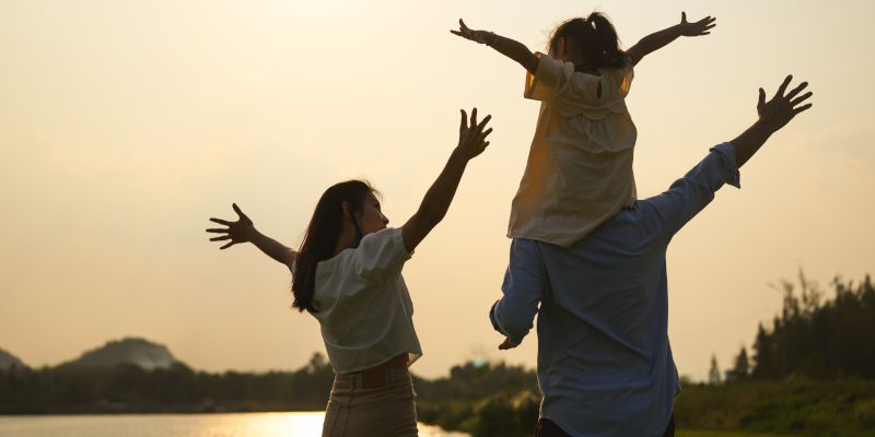 Family of three, arms outstretched, enjoying a golden sunset near water. Child sits on father's shoulders.