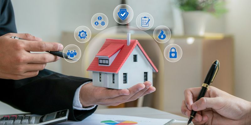 Hands holding a house model with security icons above it; another hand writing with a pen.