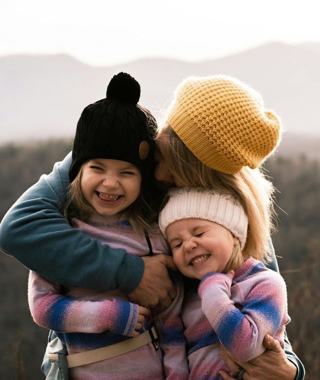 A person hugs two smiling children wearing knitted hats, with mountains in the background.