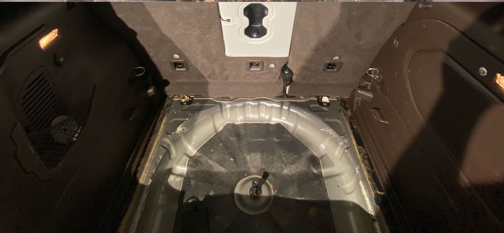 Interior of a car's trunk, showing the spare tire well and surrounding compartments. Brown and silver tones.