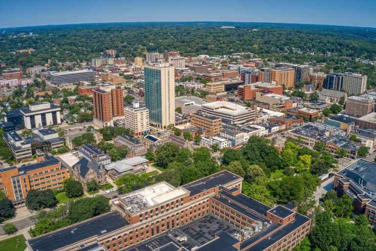 Aerial view of a city skyline with tall buildings, tree-lined streets, and a distant green horizon