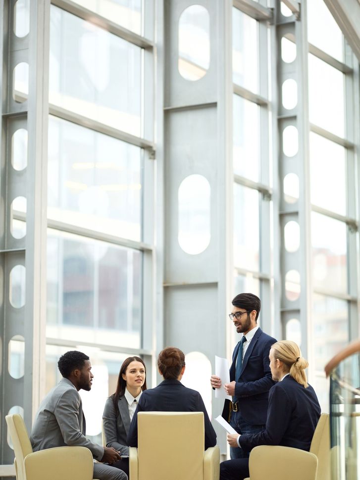 Business team meeting in a bright glass conference room, with one man standing and speaking to seated colleagues.