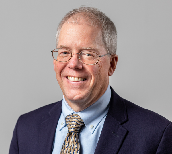 Smiling man in glasses wearing a dark blazer, blue shirt, and patterned tie against a gray background