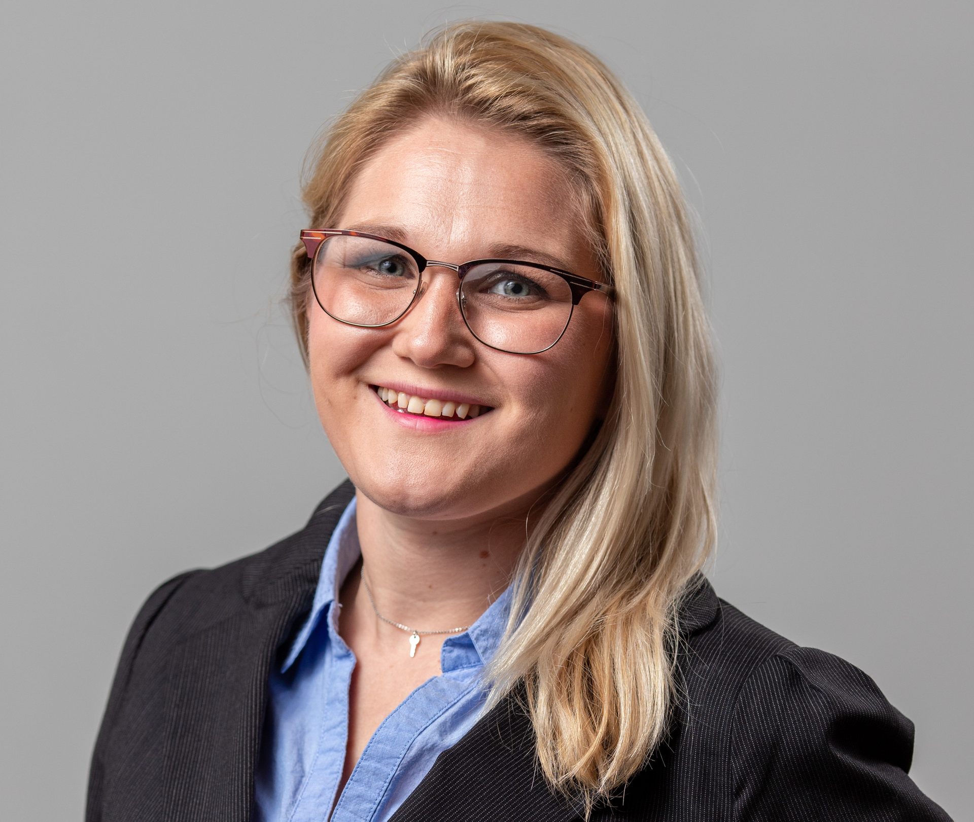 Smiling blonde woman wearing glasses and a blazer against a gray background