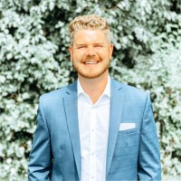 Smiling man in a blue suit and white shirt standing before leafy greenery