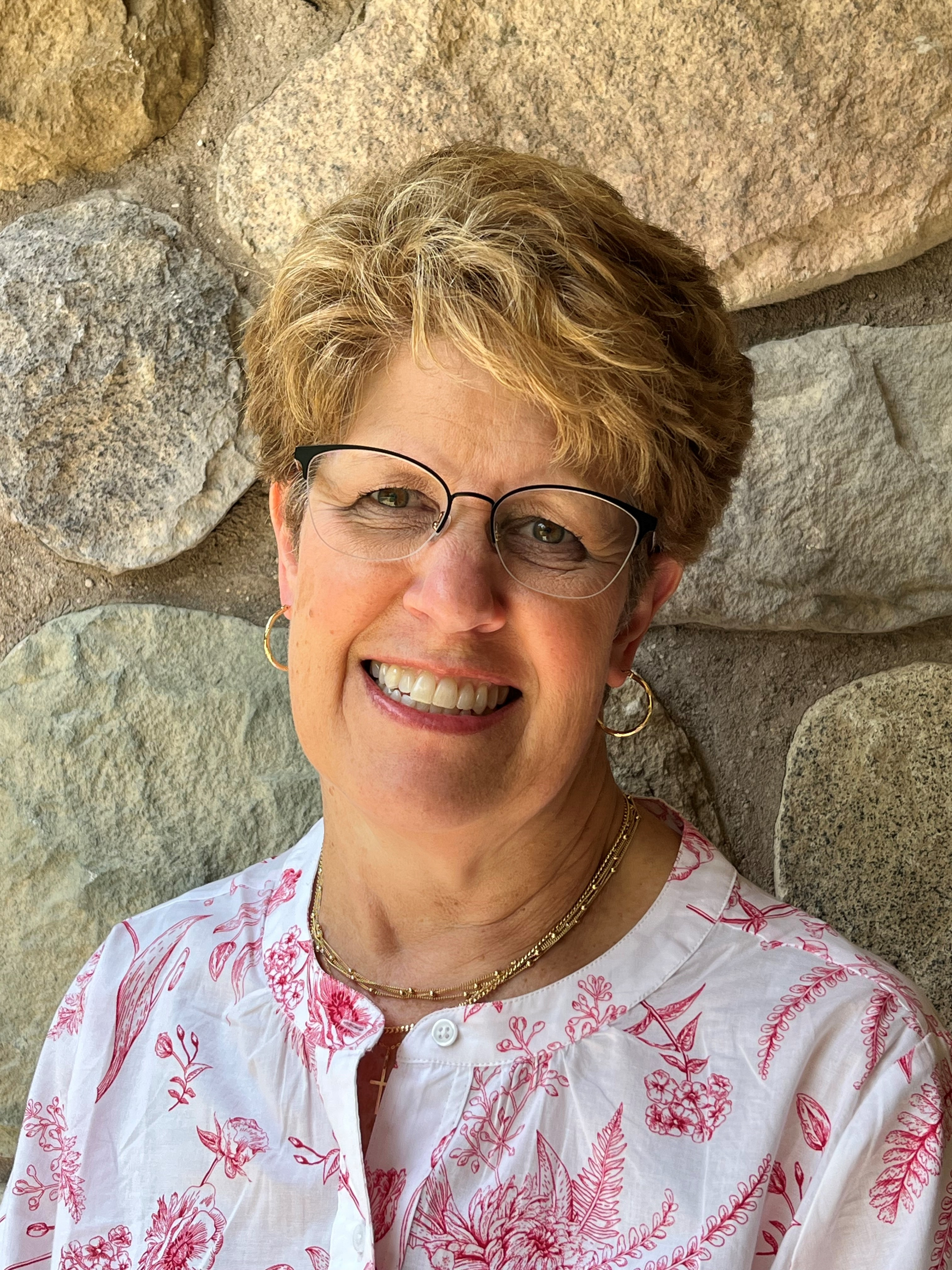 Smiling woman in glasses standing before a stone wall, wearing a white blouse with pink floral print.