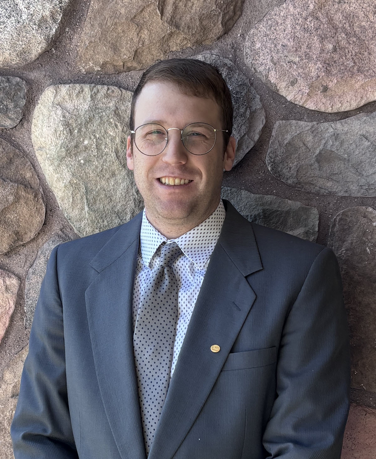 Man in suit and tie with glasses, standing in front of a stone wall and smiling.