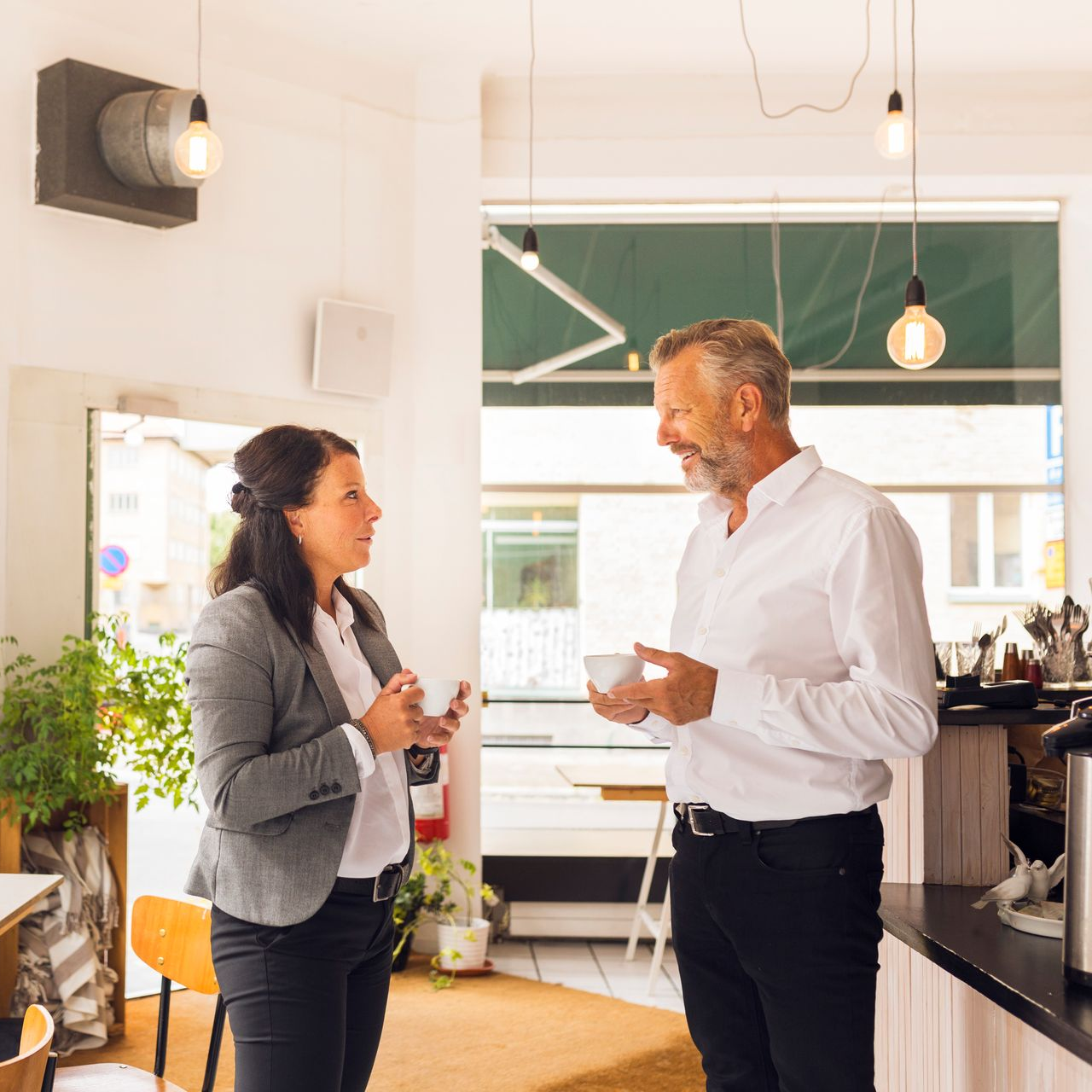 Two coworkers chatting in a bright office, holding coffee near a counter and large windows.