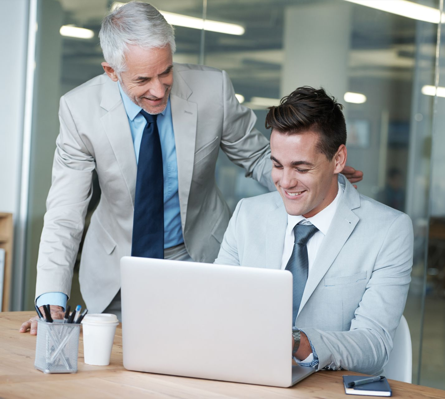 Two men in suits smiling at a laptop in a bright office.