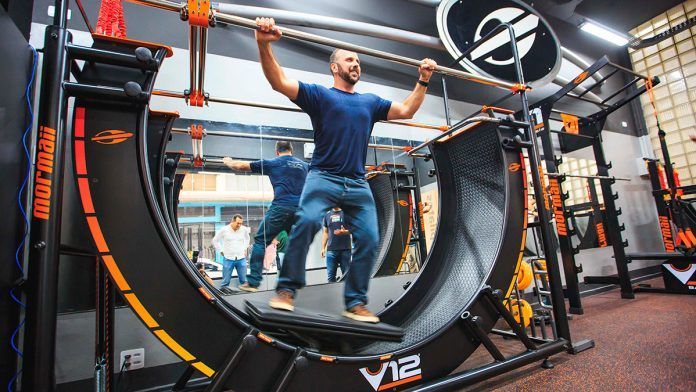 Homem se exercita em uma plataforma curva V12, segurando uma barra. Ambiente de academia com outra pessoa ao fundo.