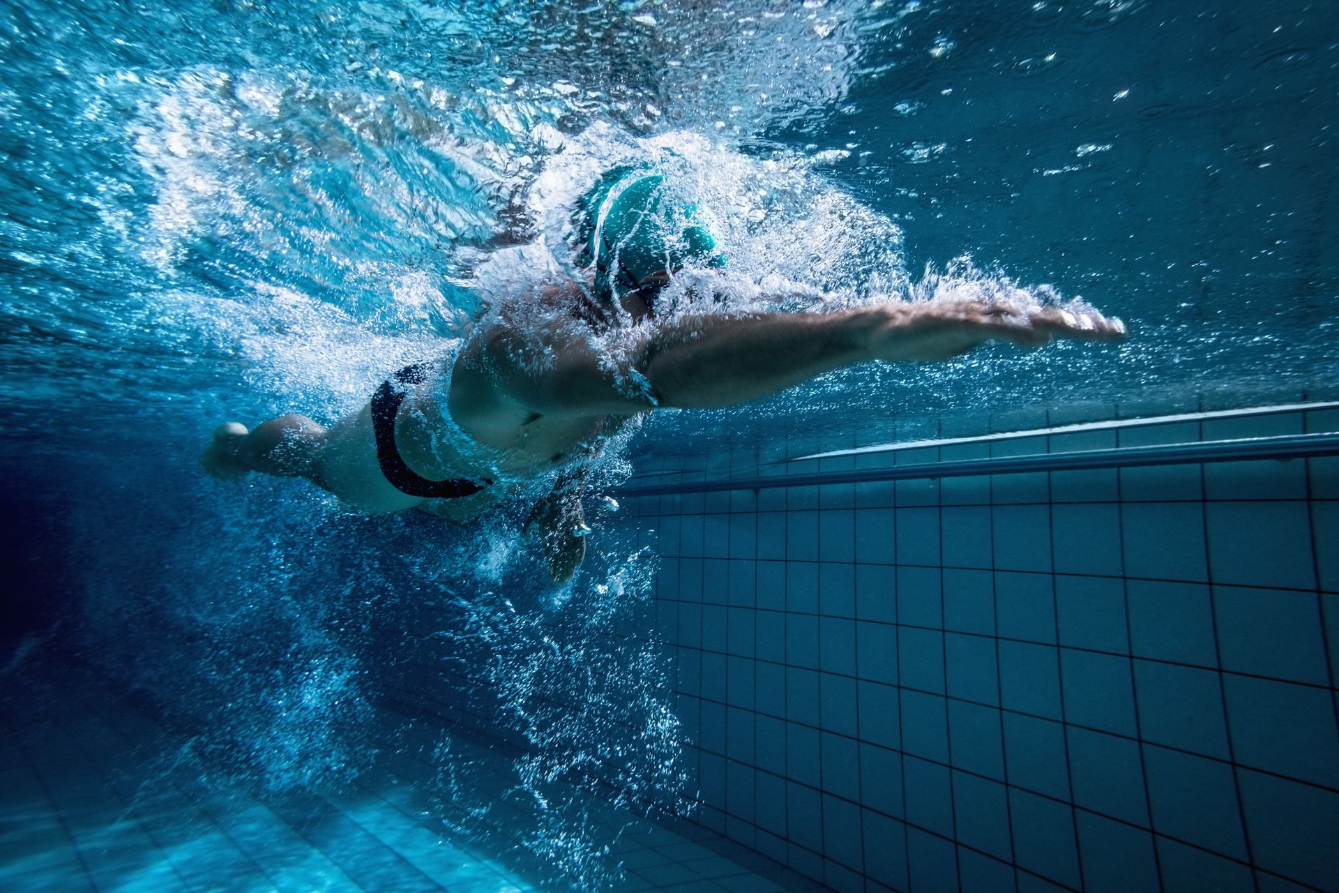 Nadador em uma piscina, submerso, braços estendidos, água azul e parede de azulejos.