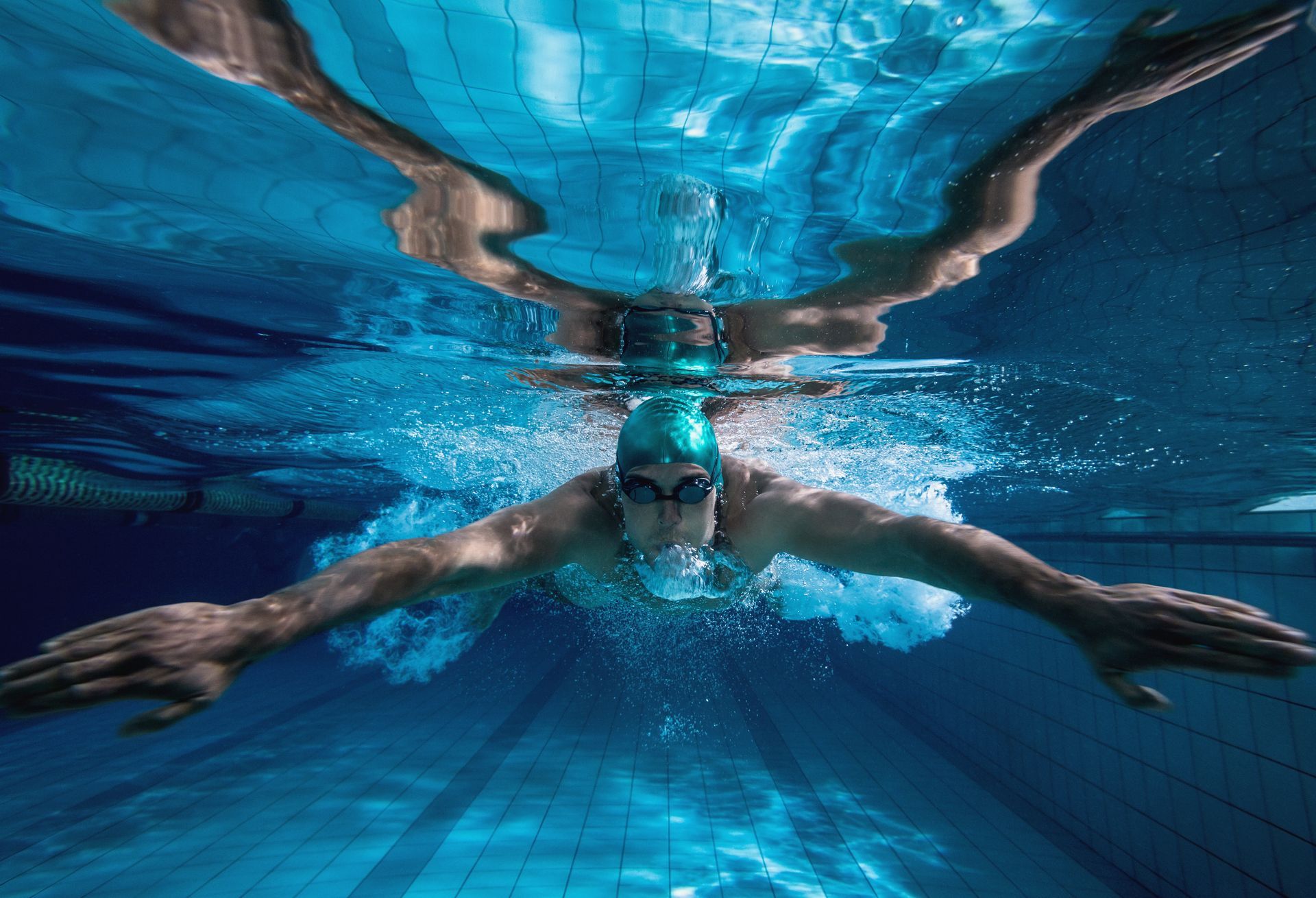 Nadador submerso, braços estendidos, usando óculos de natação e touca. Piscina com azulejos azuis visíveis.