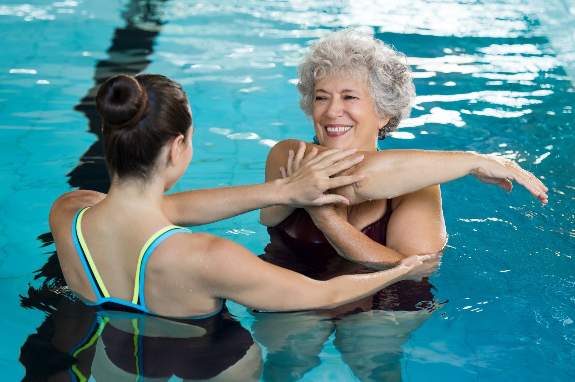 Mulher na piscina, com o braço alongado por outra mulher. Água azul, ambiente interno, sorrindo.